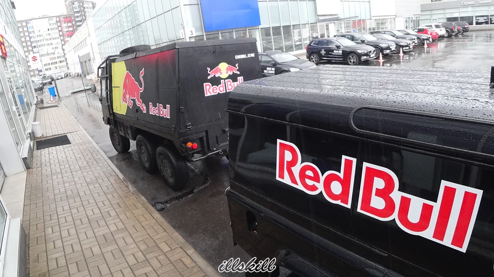 Крупный план надписи Red Bull на чёрном борту Defender; за ним виден шестиколёсный автомобиль.