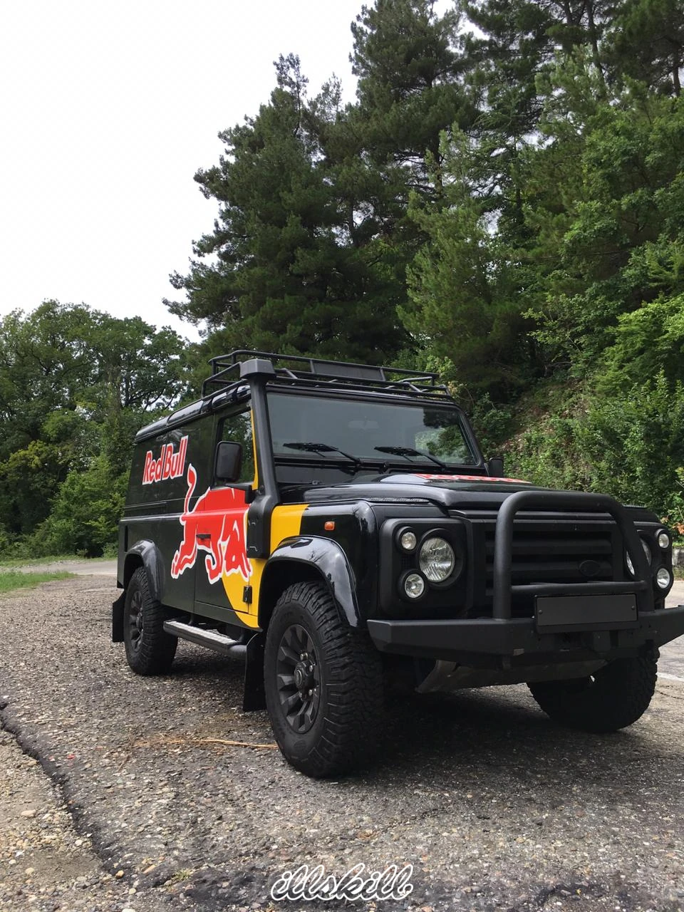 Чёрный Land Rover Defender с изображением красного быка на борту, припаркованный у горной дороги.